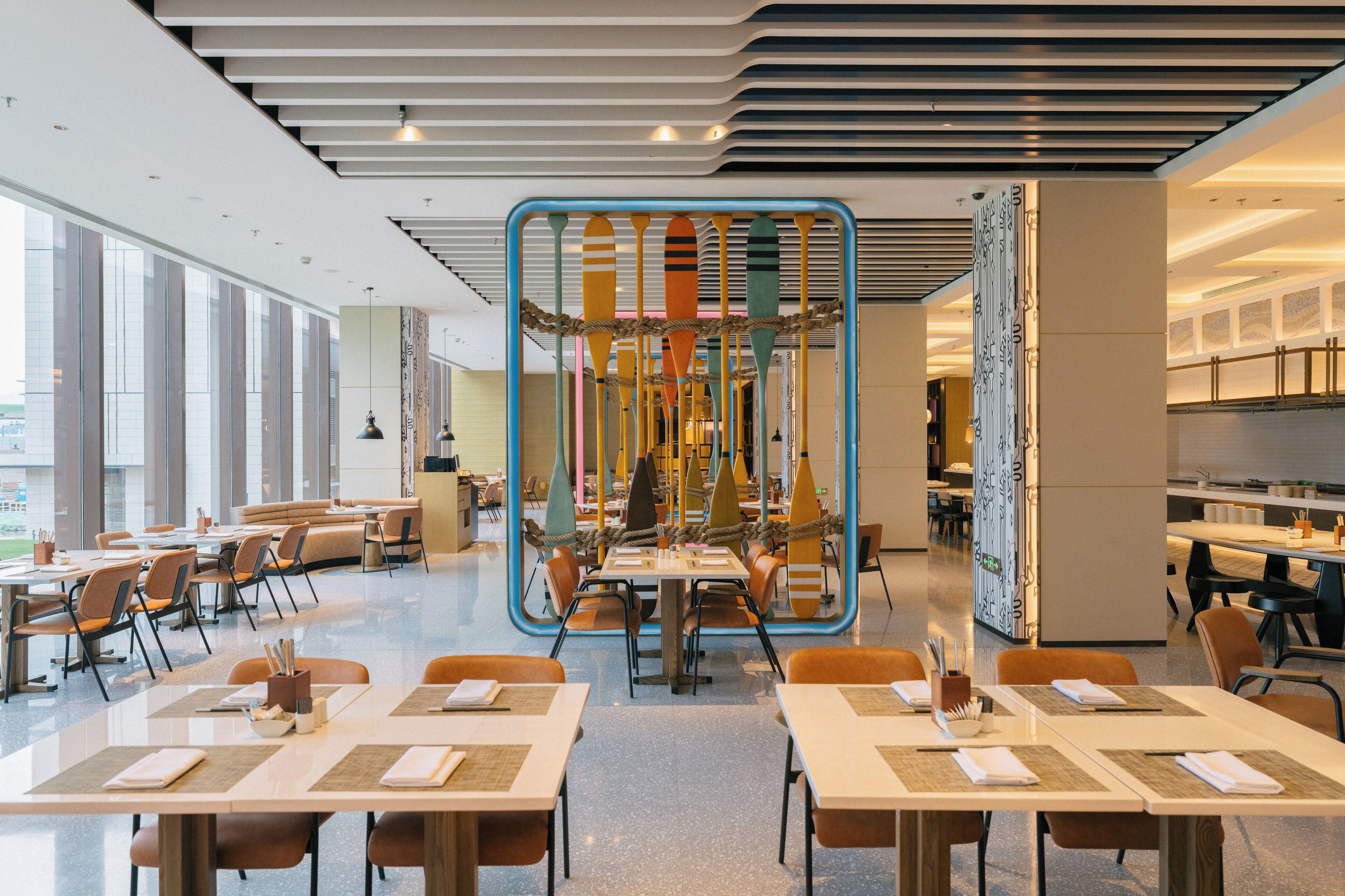 Un restaurante moderno con una zona central de mesas alrededor de una estructura decorativa única. El espacio está bien iluminado gracias a amplias ventanas que dejan entrar luz natural.