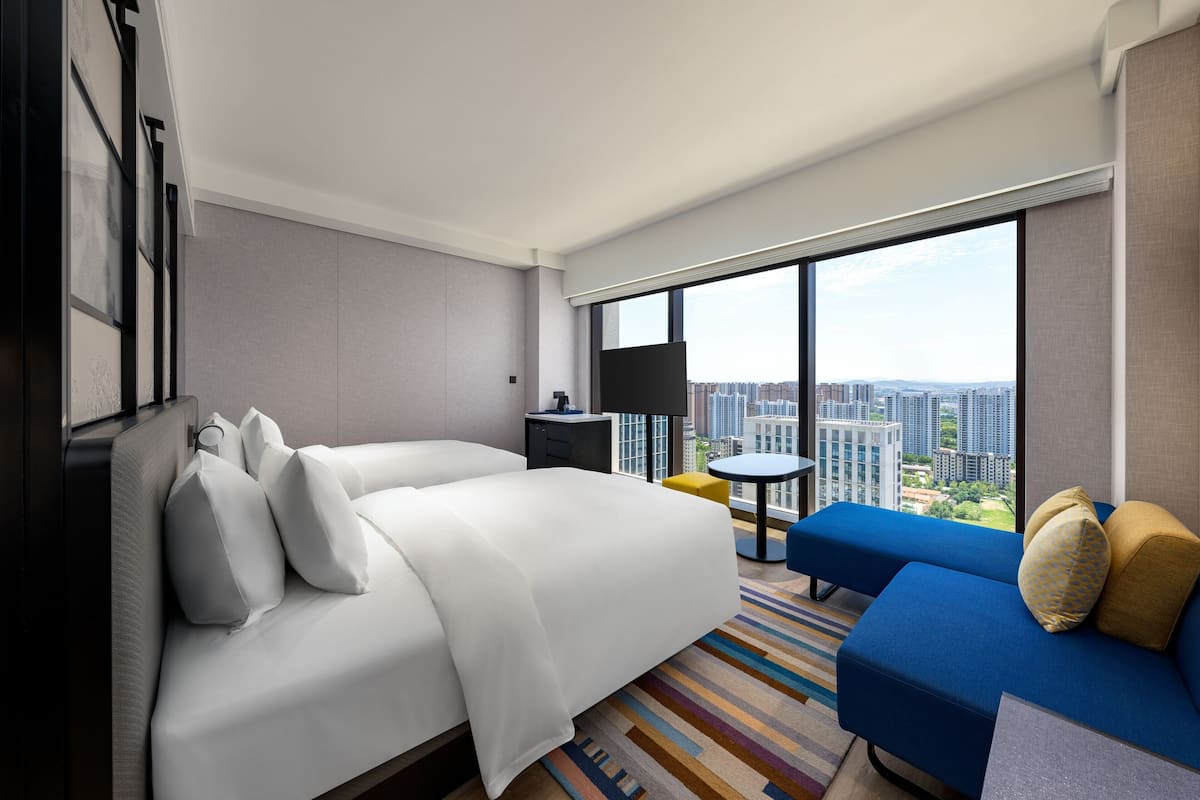 Habitación de hotel con una cama grande, un sofá azul, una mesita y vistas a la ciudad a través de amplios ventanales.