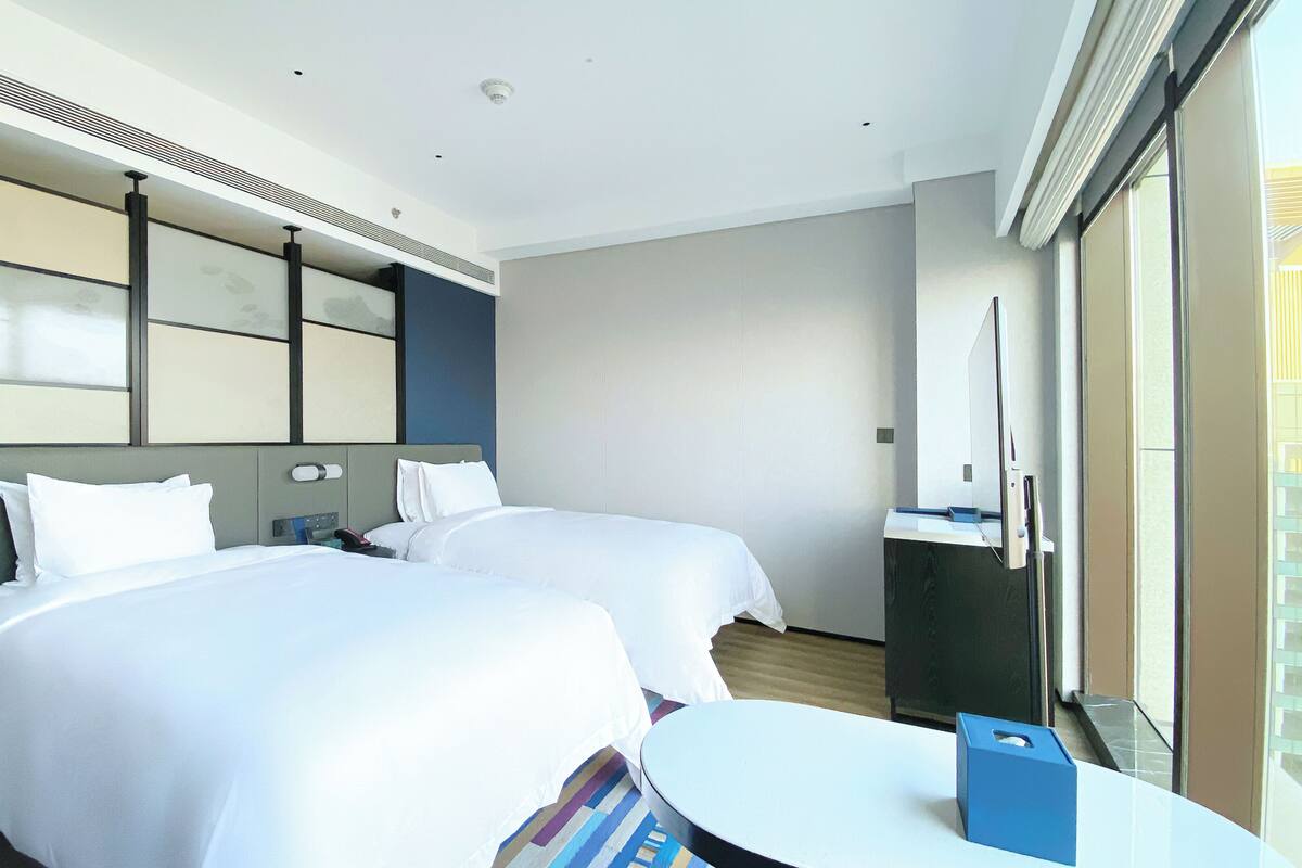 Una habitación de hotel con dos camas, una mesita pequeña y un ventanal grande.