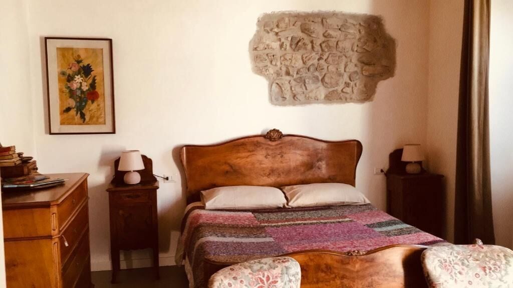 Un dormitori amb un llit de fusta, una paret de pedra, un quadre amb marc, una tauleta de nit i una làmpada.
