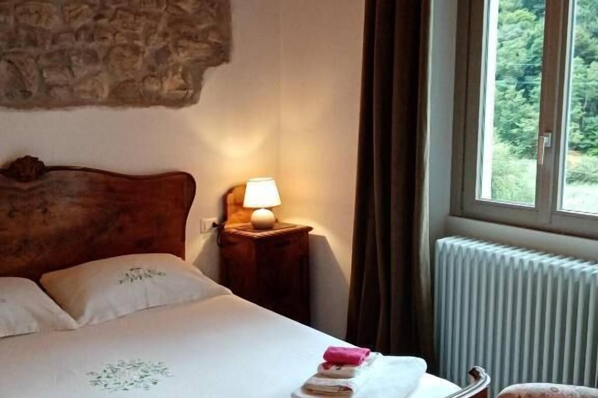 Une chambre avec un lit en bois, un mur en pierre, une table de chevet avec une lampe, un radiateur et une fenêtre donnant sur un paysage verdoyant.