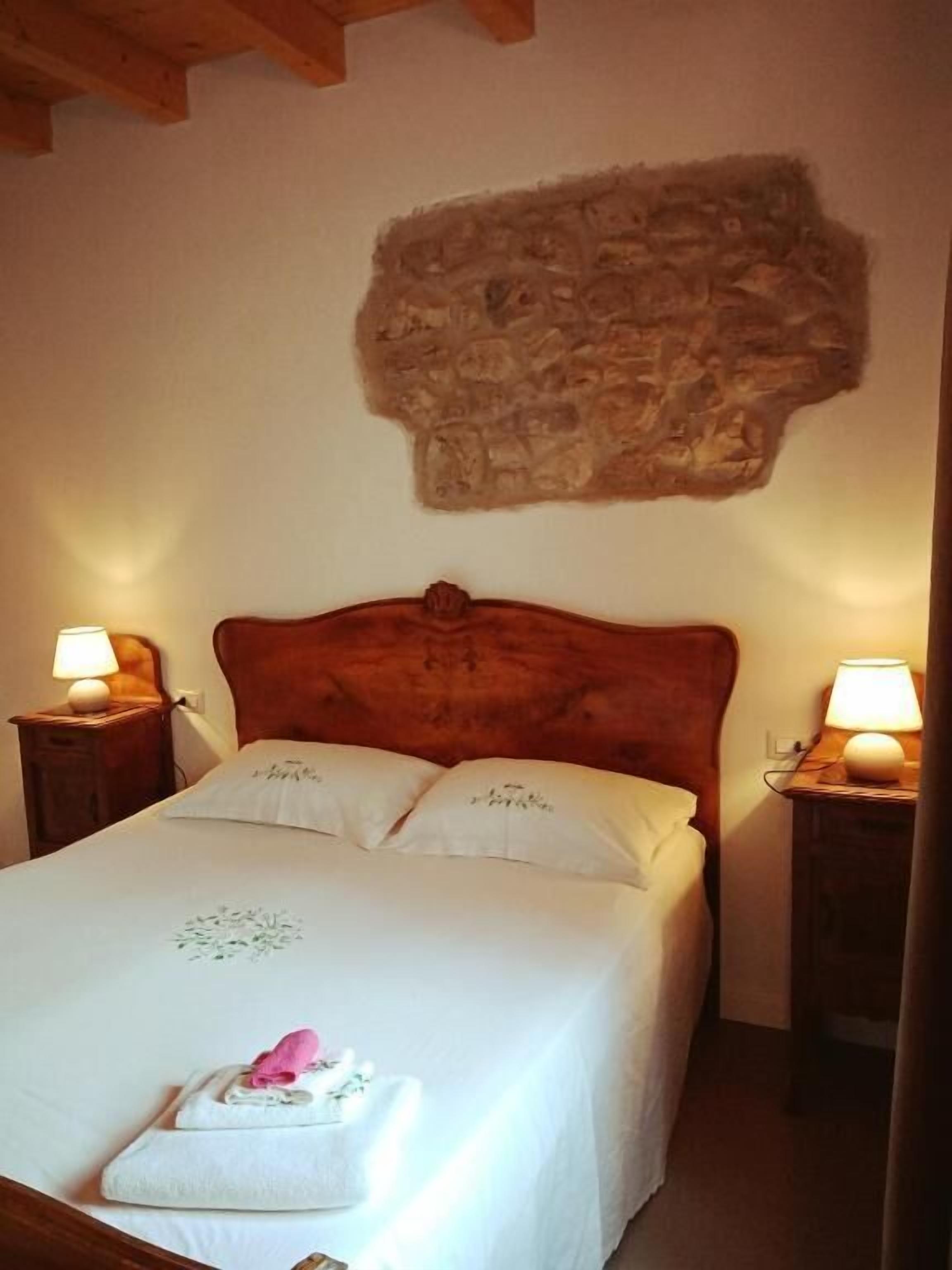 Een net opgemaakt bed met witte lakens en twee kussens, een stenen muur erboven en twee bedlampen op houten nachtkastjes.