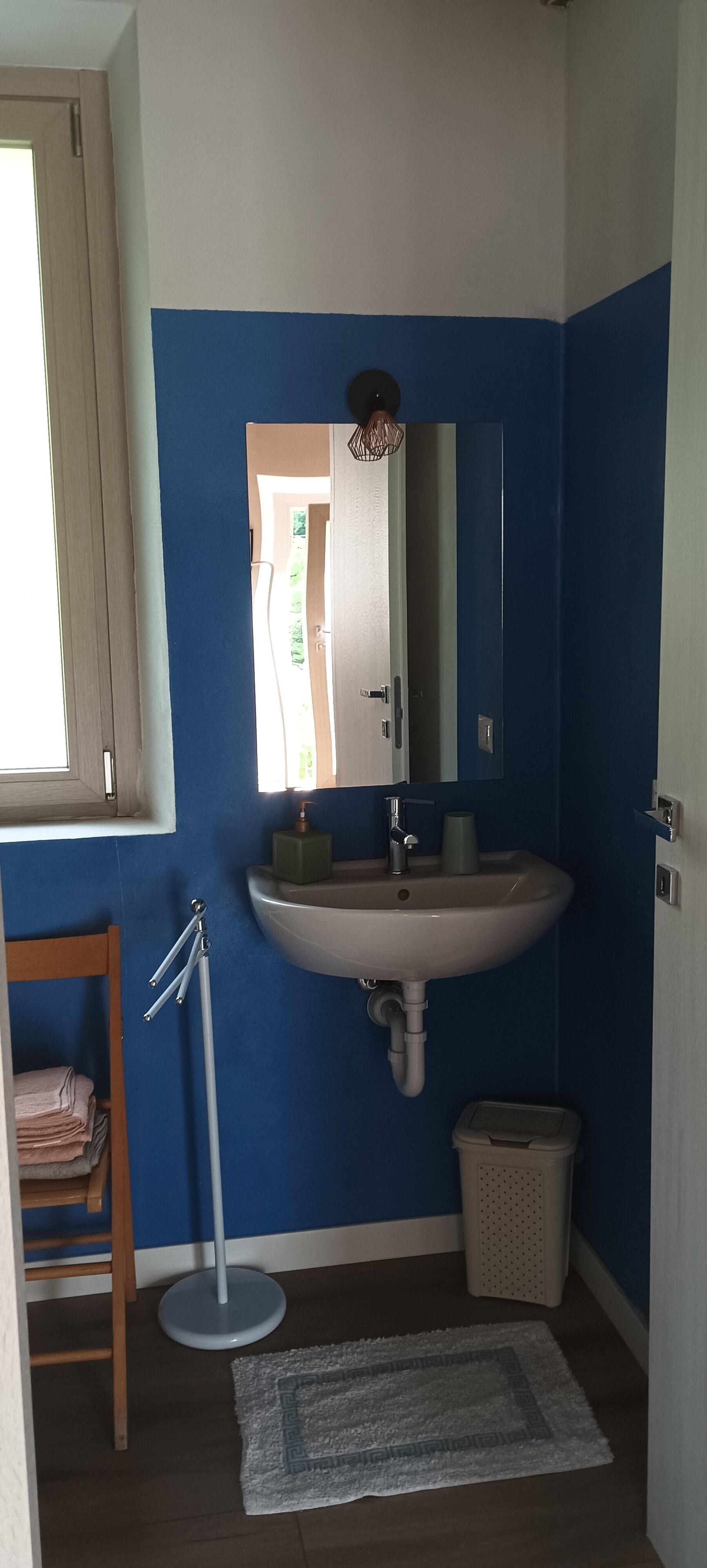 Une salle de bain avec un lavabo blanc, des murs bleus, un miroir, une chaise en bois et une petite poubelle.