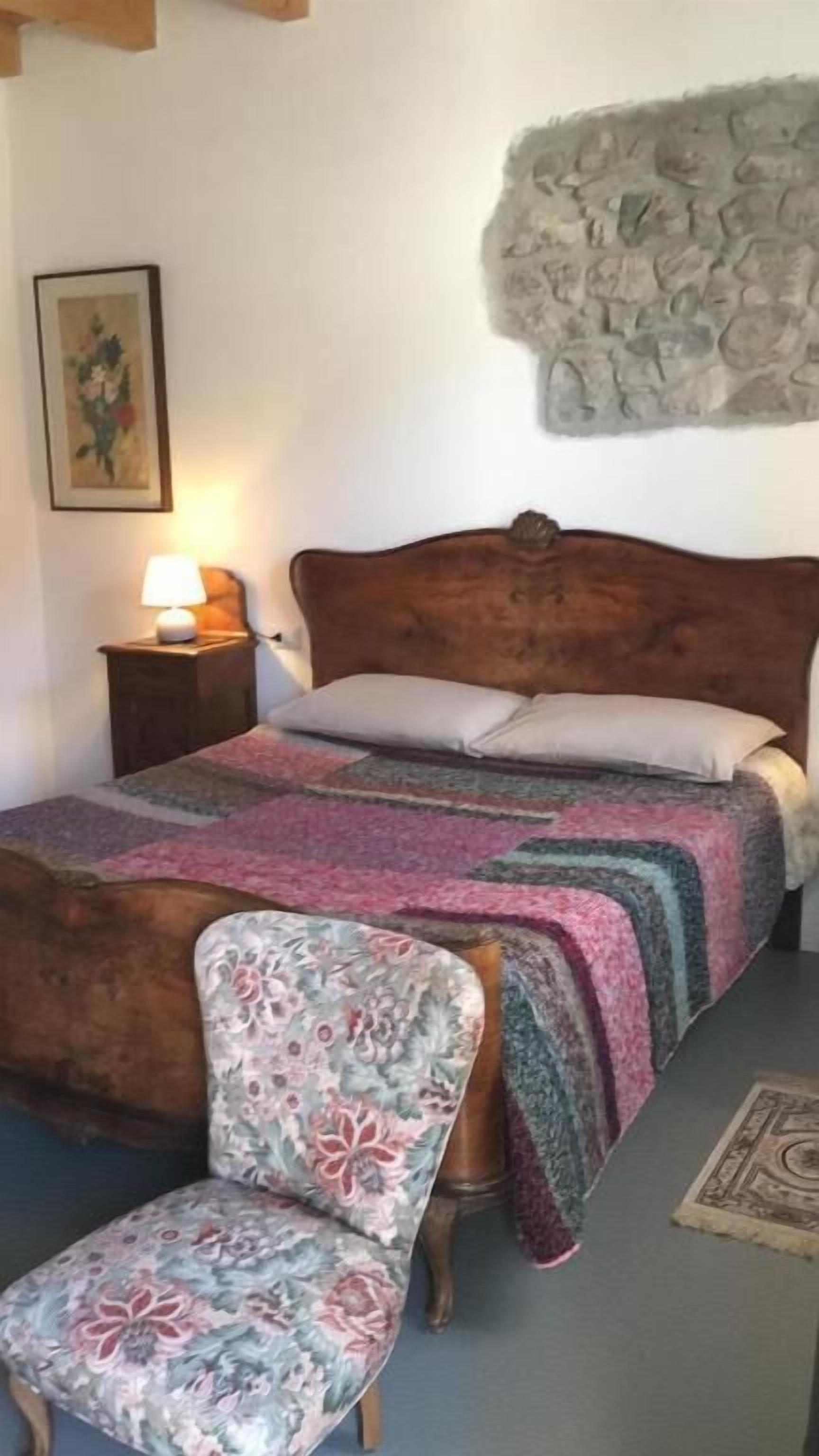 Une chambre à coucher avec un lit en bois, une chaise à motifs floraux, un mur en pierre et une table de chevet avec une lampe.