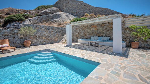 A pool area with stone flooring, a seating area with cushions, and a pergola structure.