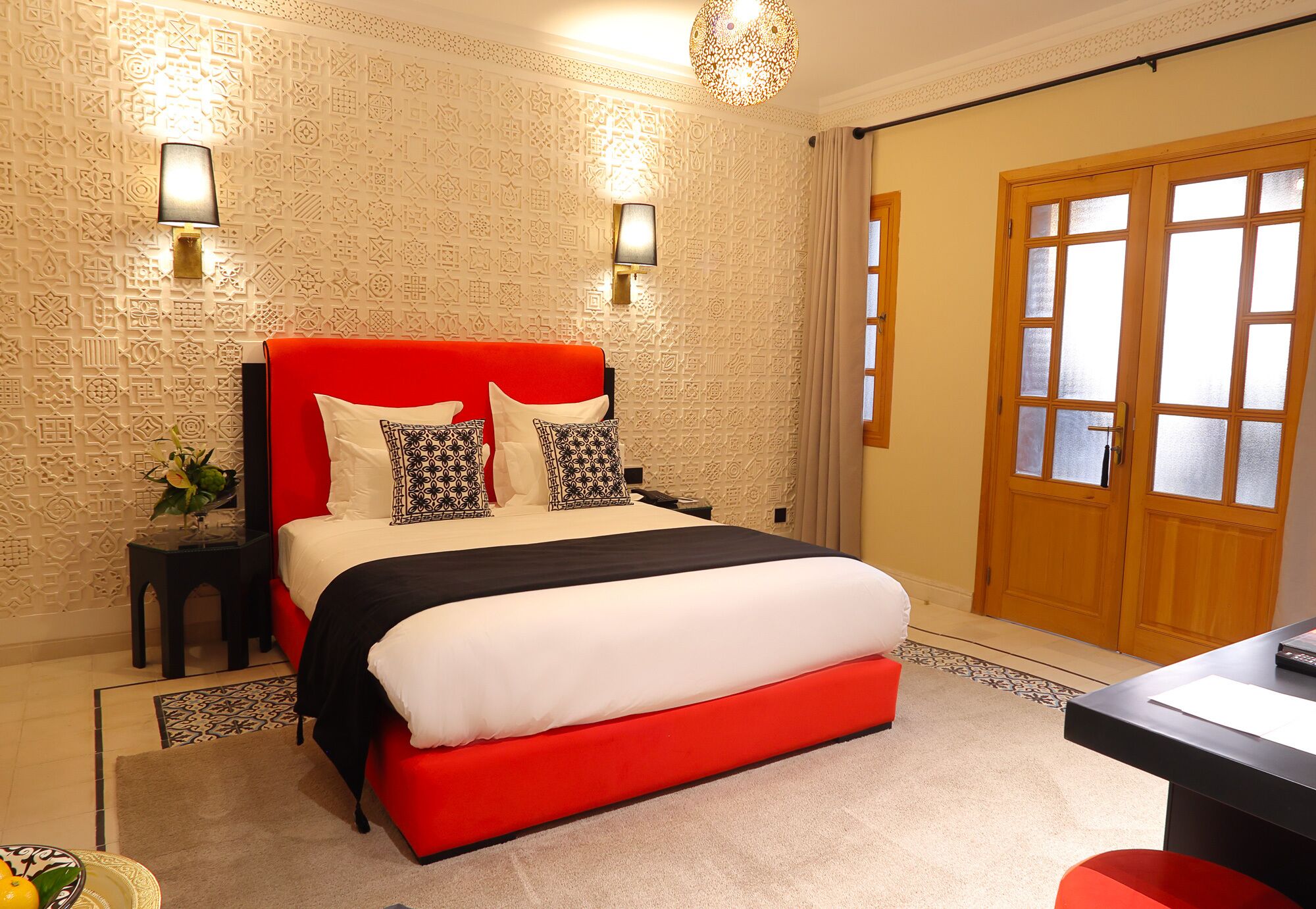 A hotel room with a red bed, patterned wallpaper, two wall-mounted lamps, and a wooden door.