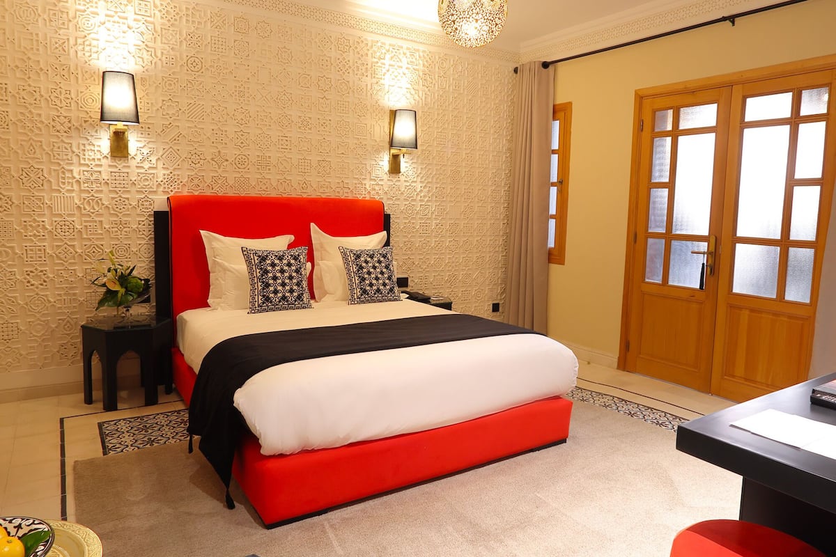 Habitación de hotel con cama roja, papel tapiz estampado, dos lámparas fijadas a la pared y una puerta de madera.