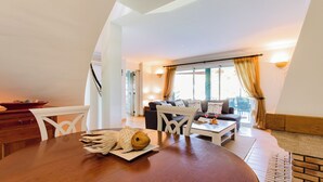 A modern living room with a wooden dining table, a sofa, and a view of the outdoors through large windows.