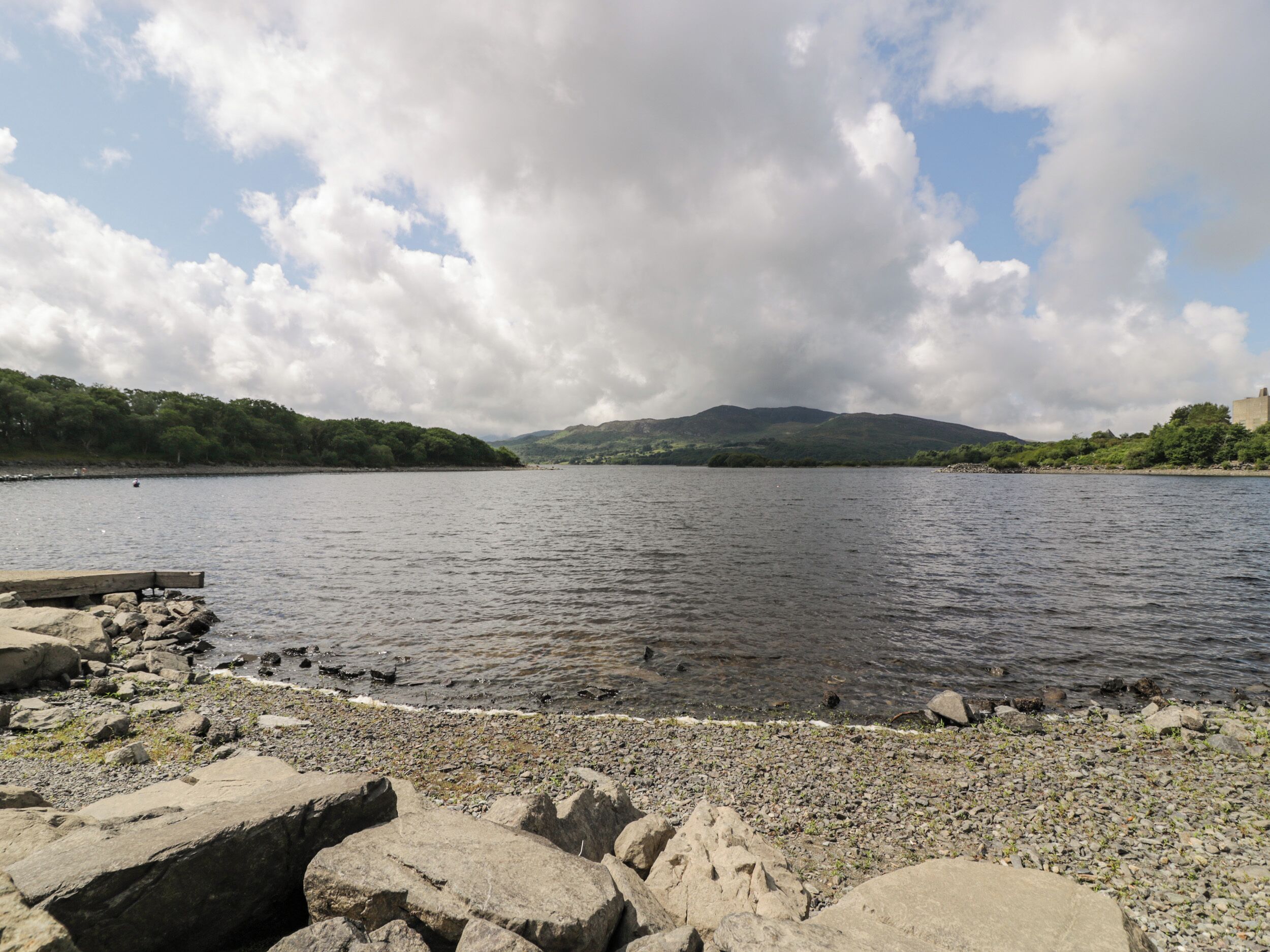 A rocky shoreline with a calm body of water, distant hills, and a partly cloudy sky.