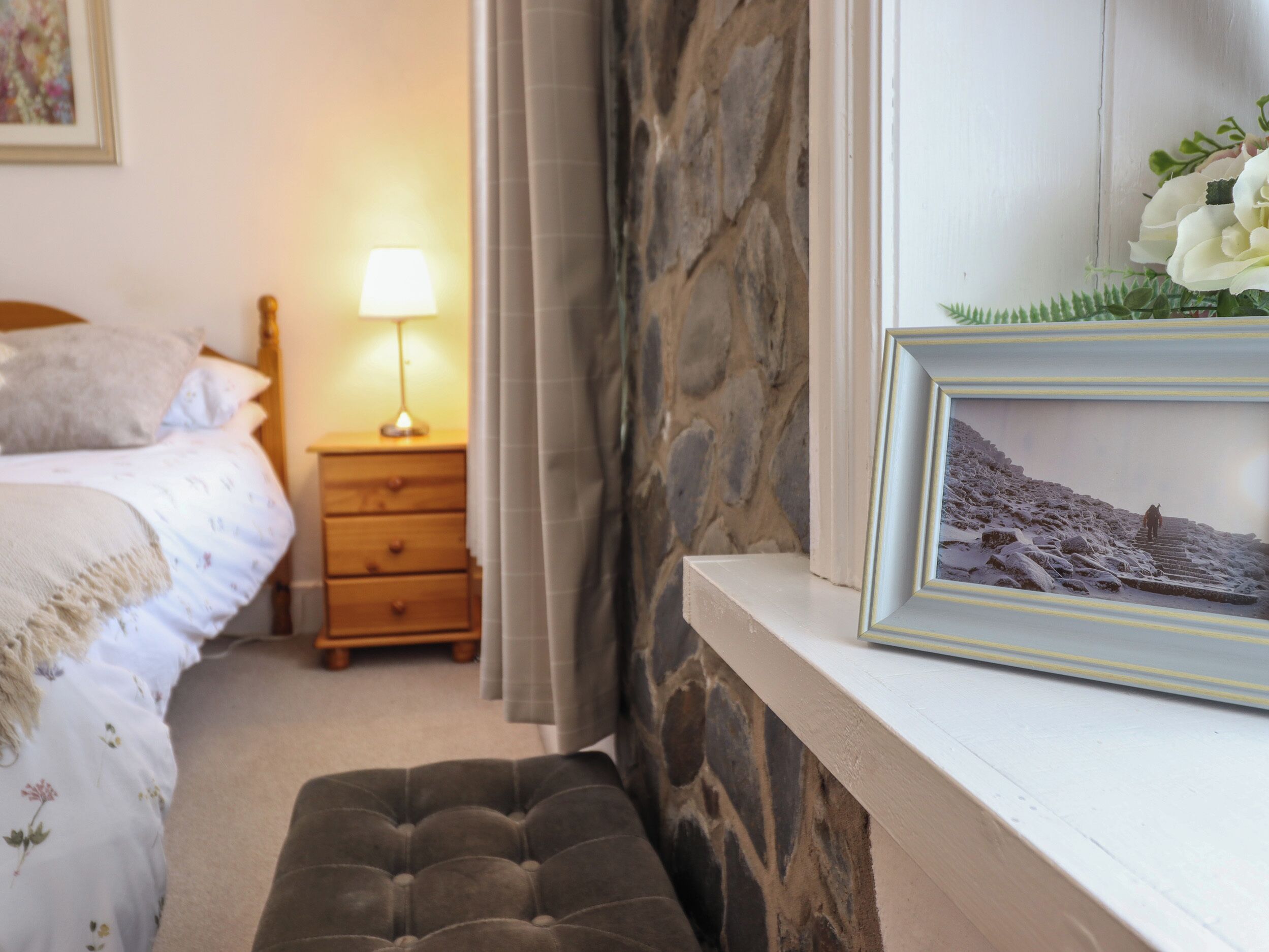 A bedroom with a bed, a nightstand with a lamp, a stone wall, and a framed picture on the dresser.