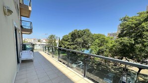 A balcony with a glass railing, a white chair, and a view of parked cars and trees. - Ameijeira Lagos Apartment (Lagos)