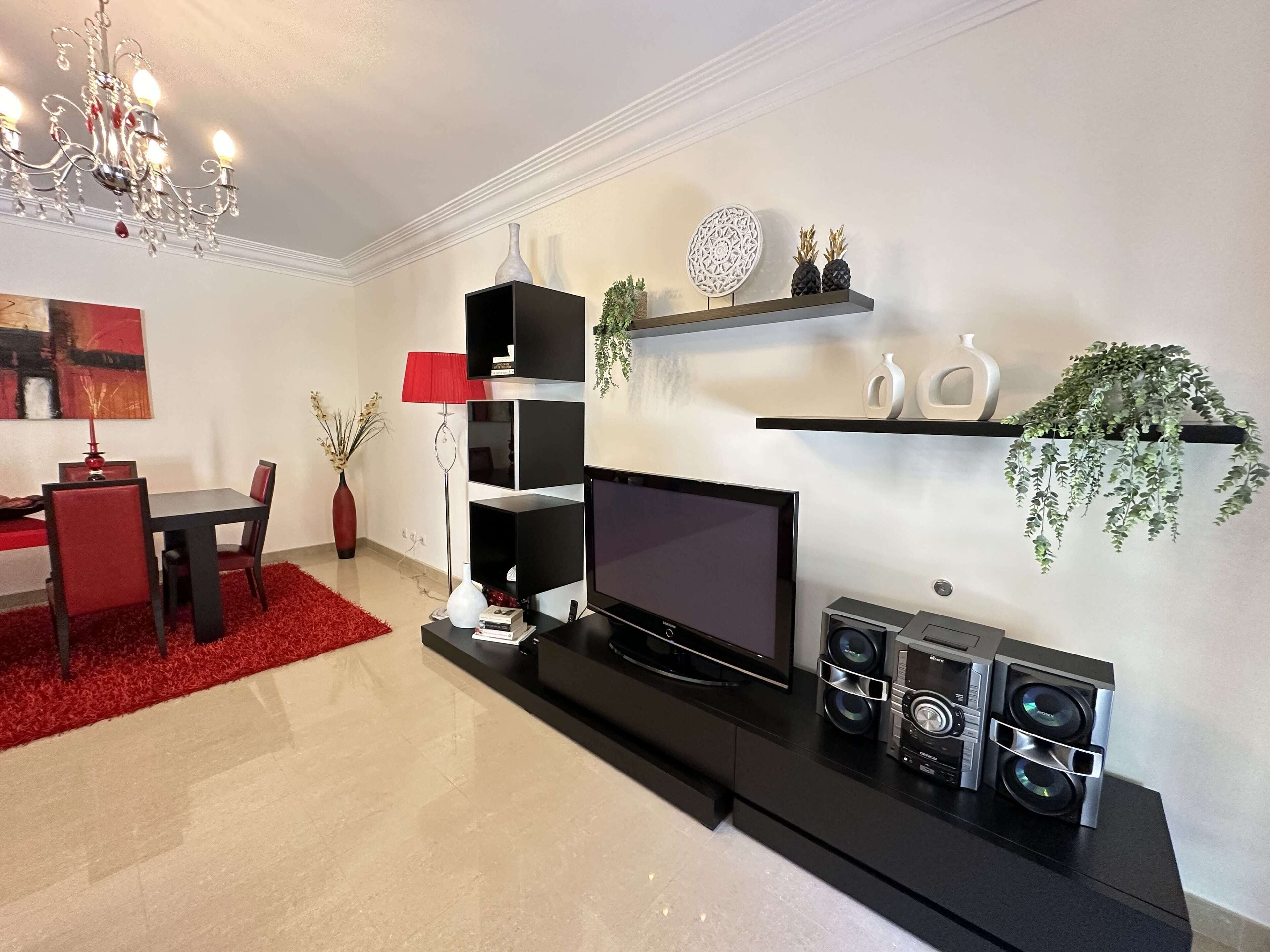 A modern living room with a flat-screen TV, black entertainment unit, red chairs, and a red rug.