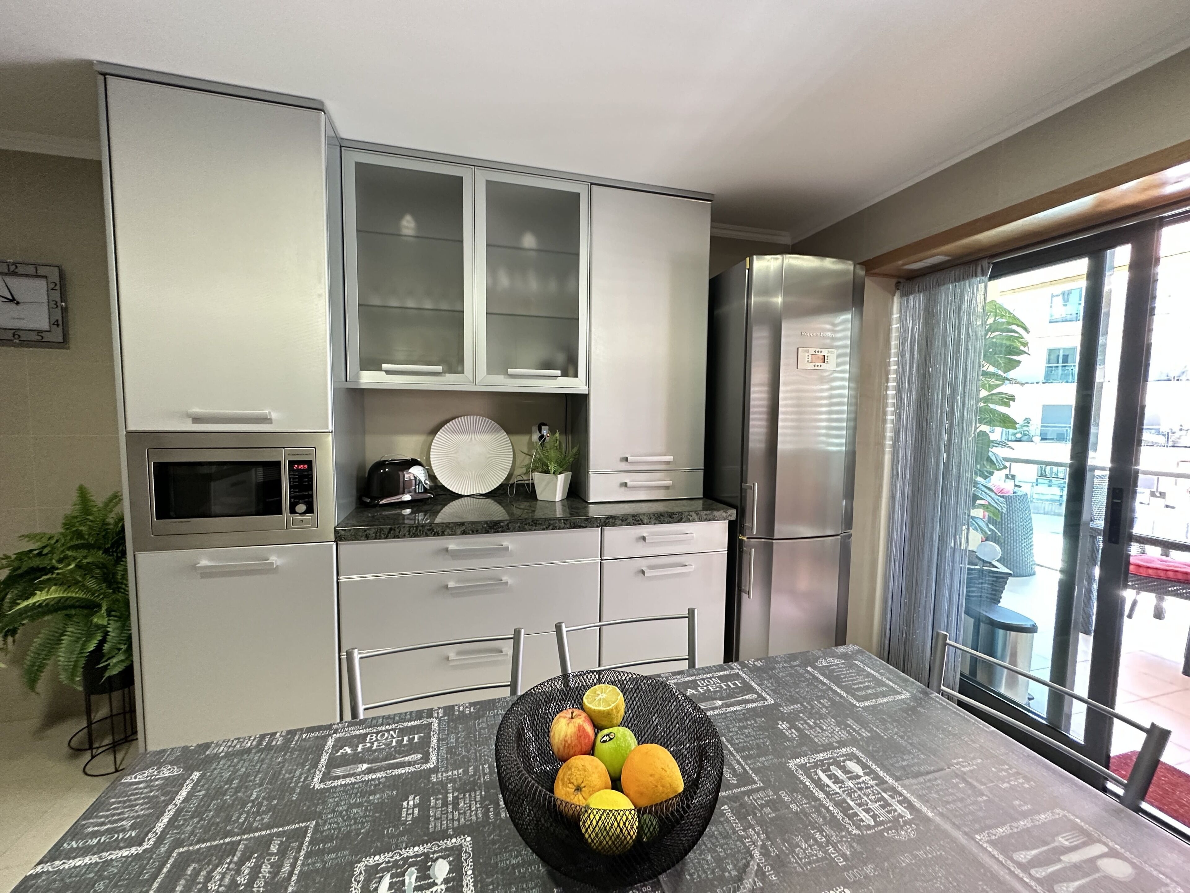 A modern kitchen with stainless steel appliances, a microwave, and a built-in oven. There is a dining table with a black and white patterned tablecloth and a fruit basket on it.