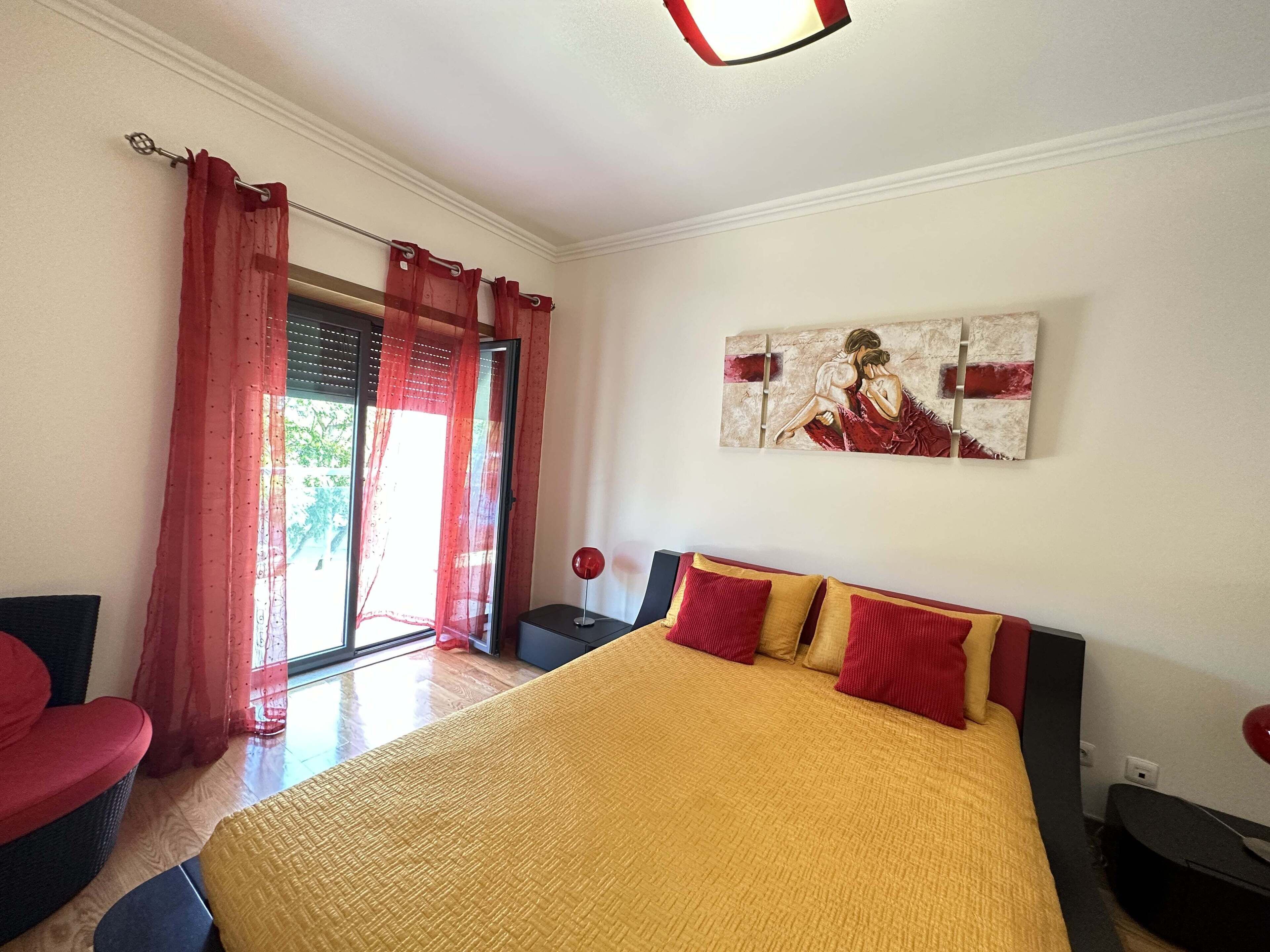 A bedroom with a large bed, red curtains, a painting on the wall, and a view through the window.