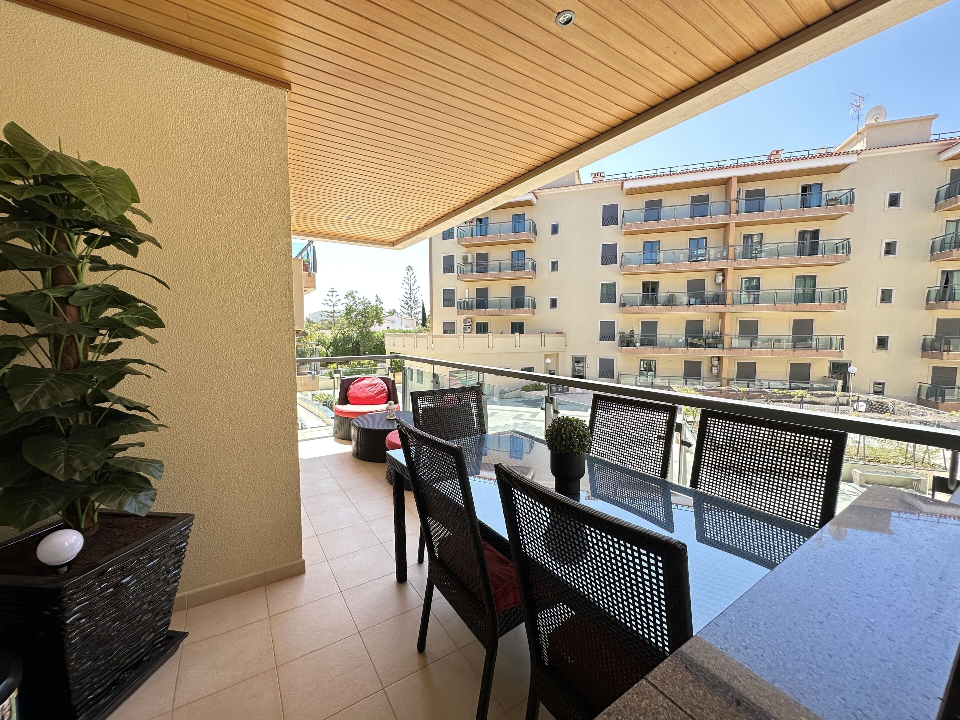 A balcony with a glass table and chairs, a potted plant, and a view of a multi-story building with balconies.
