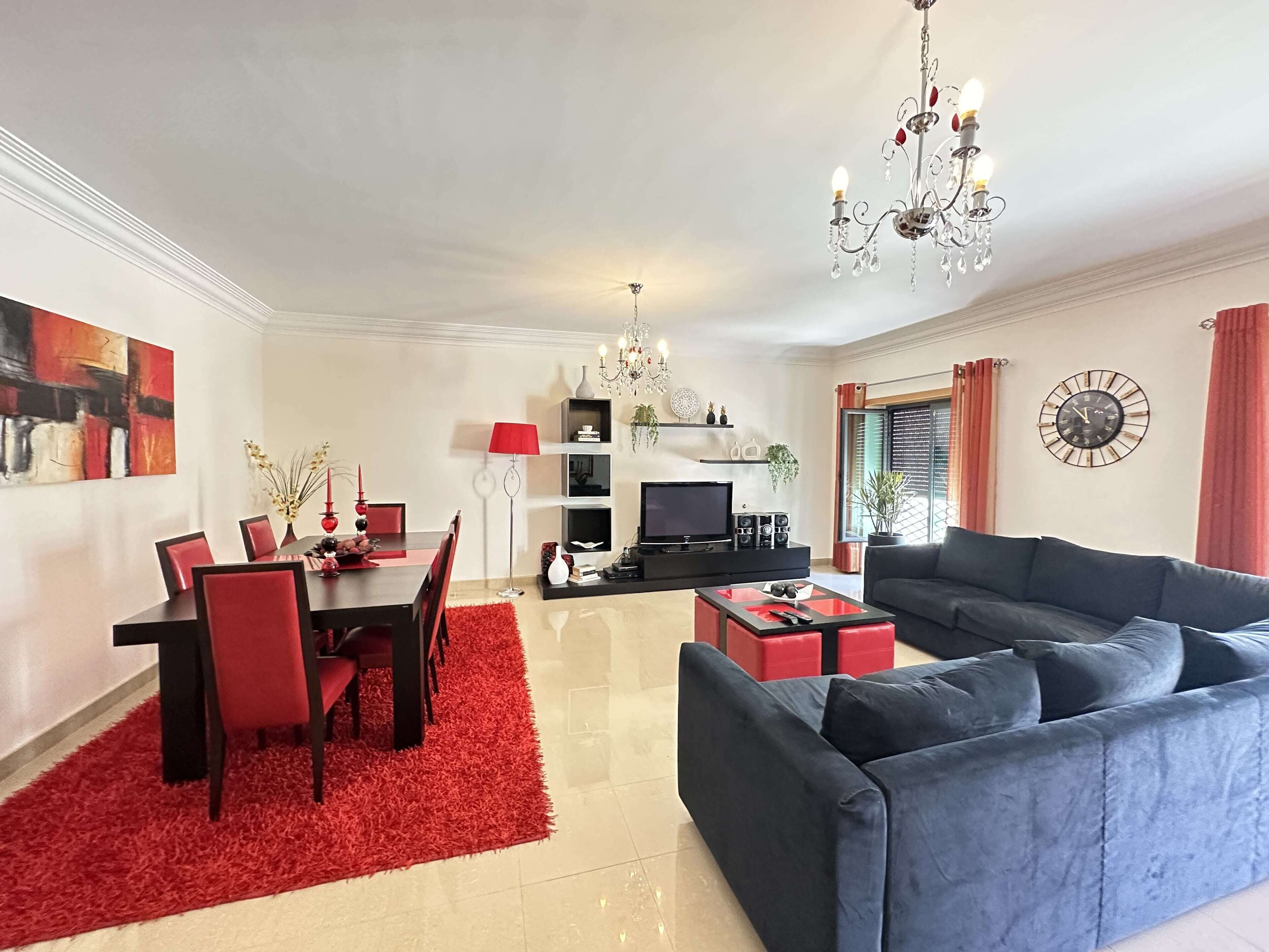 A modern living room with a blue sofa, a dining area with red chairs, a TV, and a chandelier.