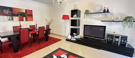 A modern living room with a dining area, a chandelier, a flat-screen TV, and a sound system.