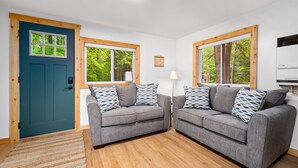 A modern living room with two grey sofas, a blue door, and large windows with a view of greenery. - Cozy l Hot Tub+Sauna l Arbutus Lake l Close to TC (Traverse City)