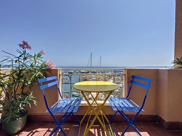 A balcony with a yellow table and blue chairs, overlooking a marina with boats.
