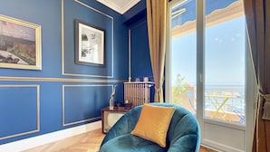 A room with a blue wall, a framed picture, a blue chair with a yellow cushion, a radiator, and a view of the sea through a balcony. - (102) Le superbe au Palais (Vallauris)