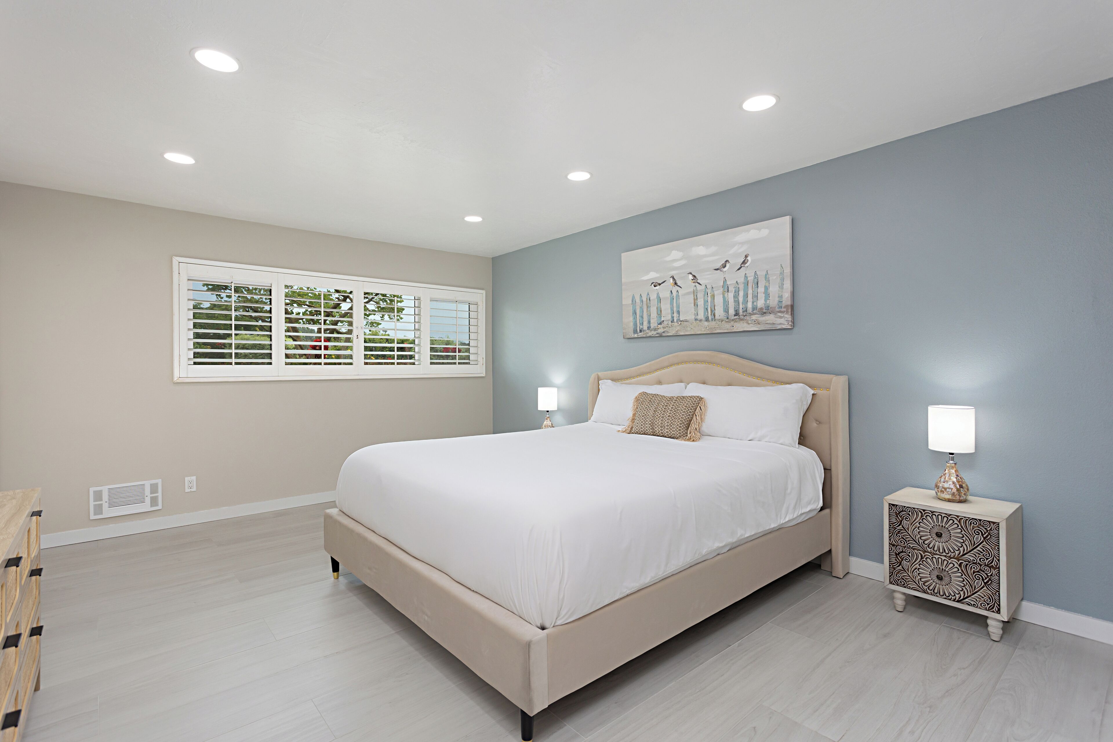A bedroom with a large bed, two bedside tables with lamps, a window with blinds, and a painting on the wall.