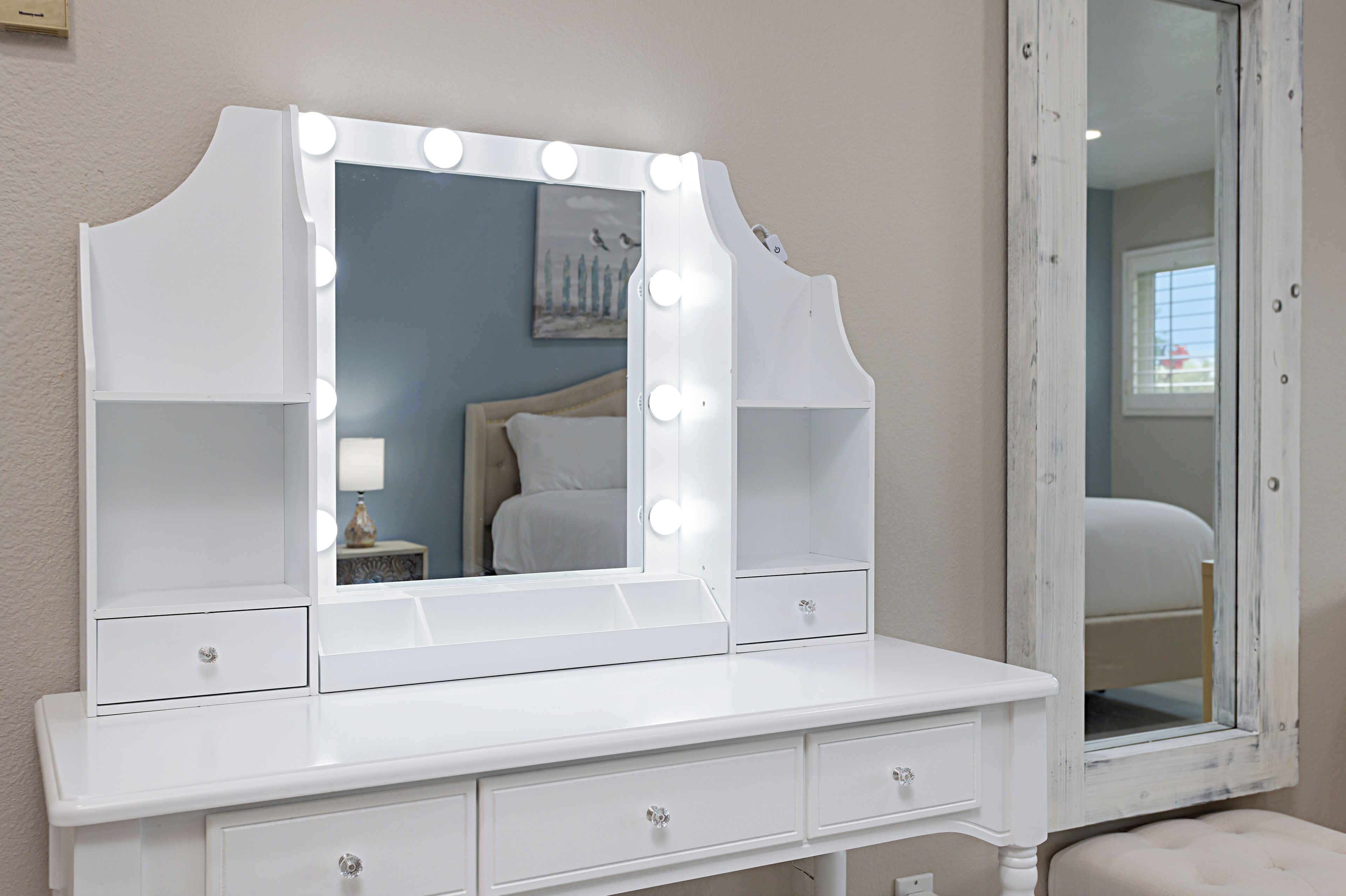 A white vanity with a large mirror and lights, a bedside table with a lamp, and a window with blinds.