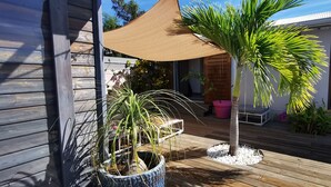 Une terrasse en bois agrémentée de plantes en pot, d’un petit palmier et d’une voile d’ombrage de couleur sable.