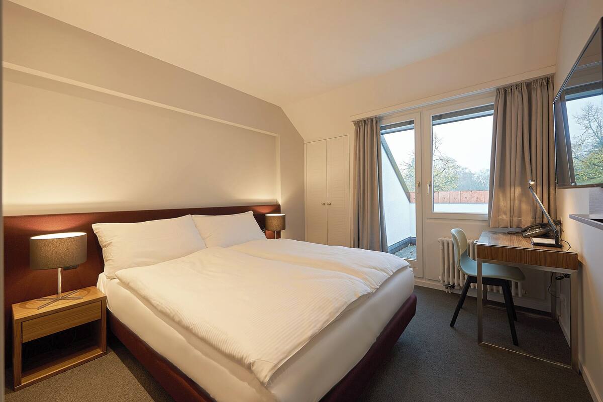 Een hotelkamer met een bed, nachtkastje, lamp, bureau, stoel en uitzicht.