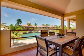 A balcony with a wooden table and chairs, overlooking a pool and resort buildings. - Magnificent Belize Townhouse-Pool-Private Island (Placencia)