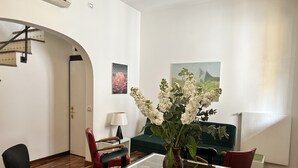 A dining area with a glass-top table, chairs, a vase with flowers, a lamp, and a painting on the wall.