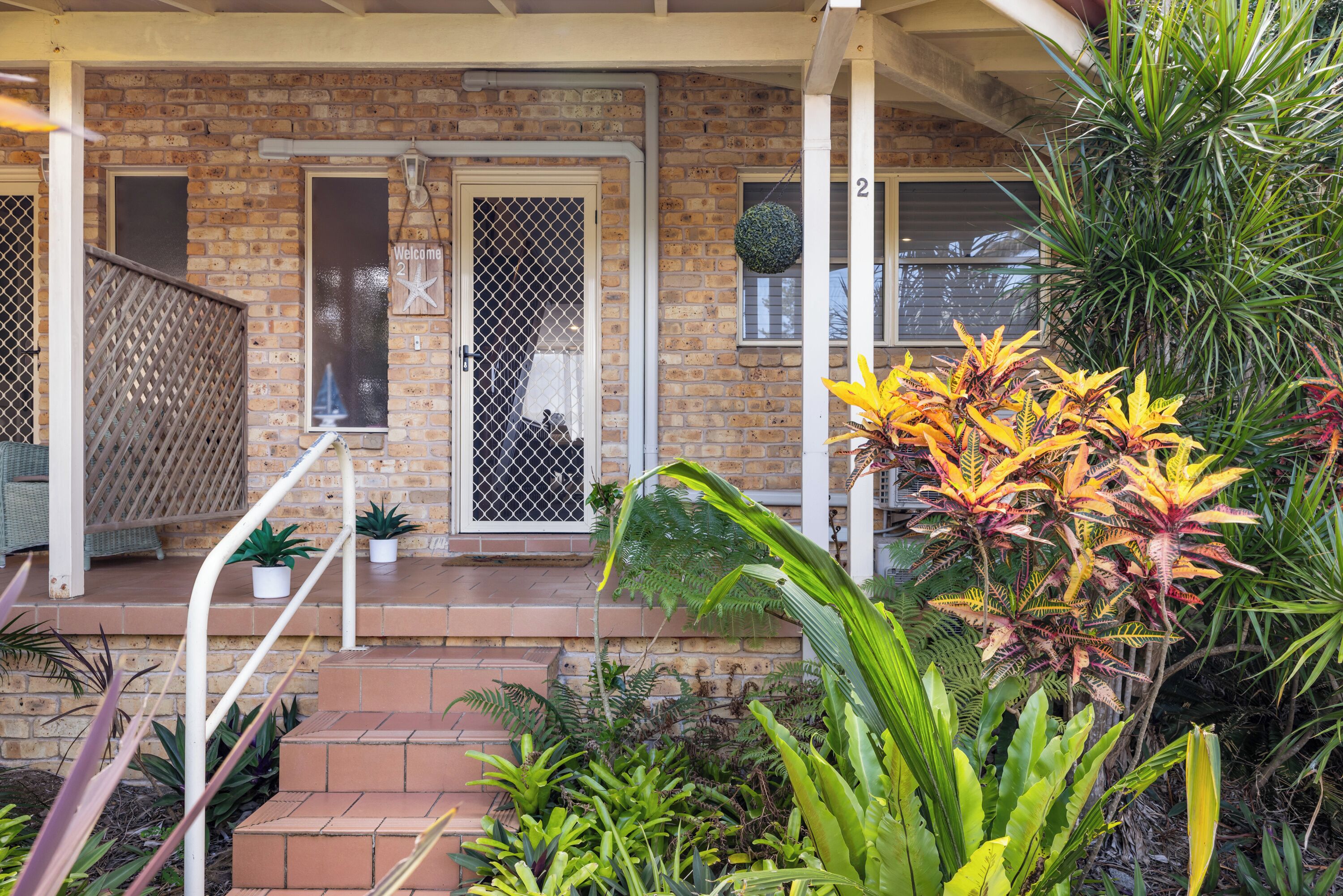 A brick house with a porch, steps, and a variety of plants.