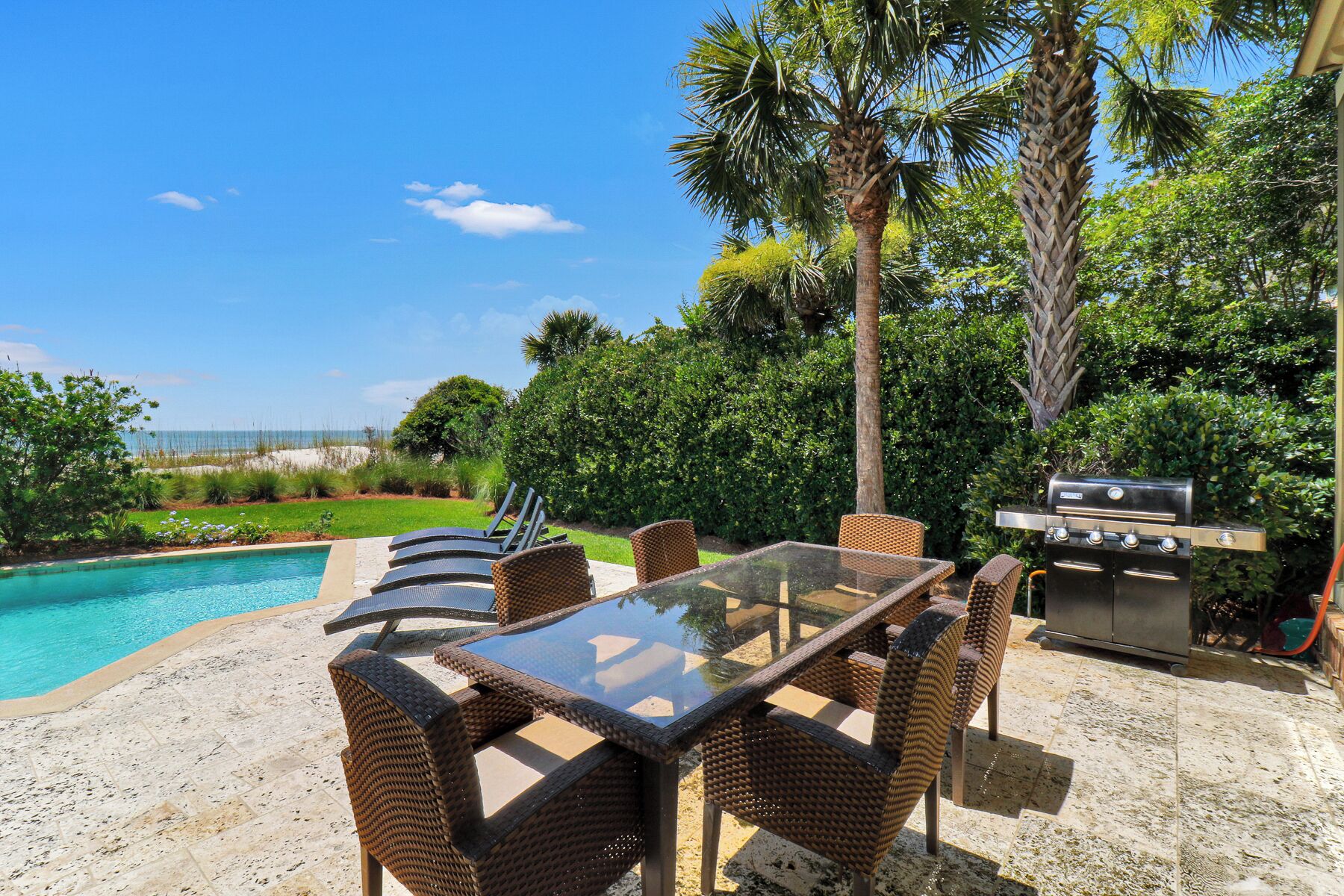 Un espace aménagé au bord de la piscine, avec une table en verre et des fauteuils en osier, un barbecue et une vue sur l’océan.