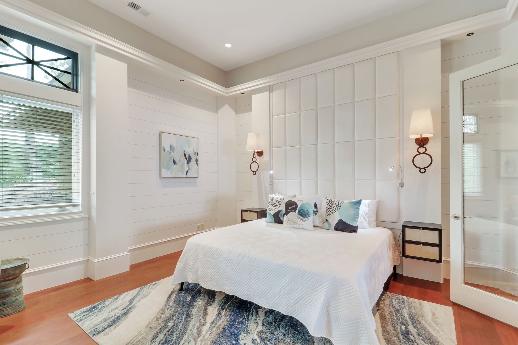 Un dormitorio con una cama grande, una ventana con persianas, un cuadro en la pared y dos lámparas fijadas a la pared.