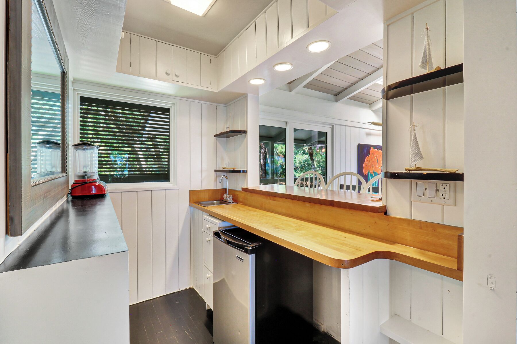 Una cocina compacta con armarios blancos, encimera de madera, lavavajillas integrado y ventana con persianas.