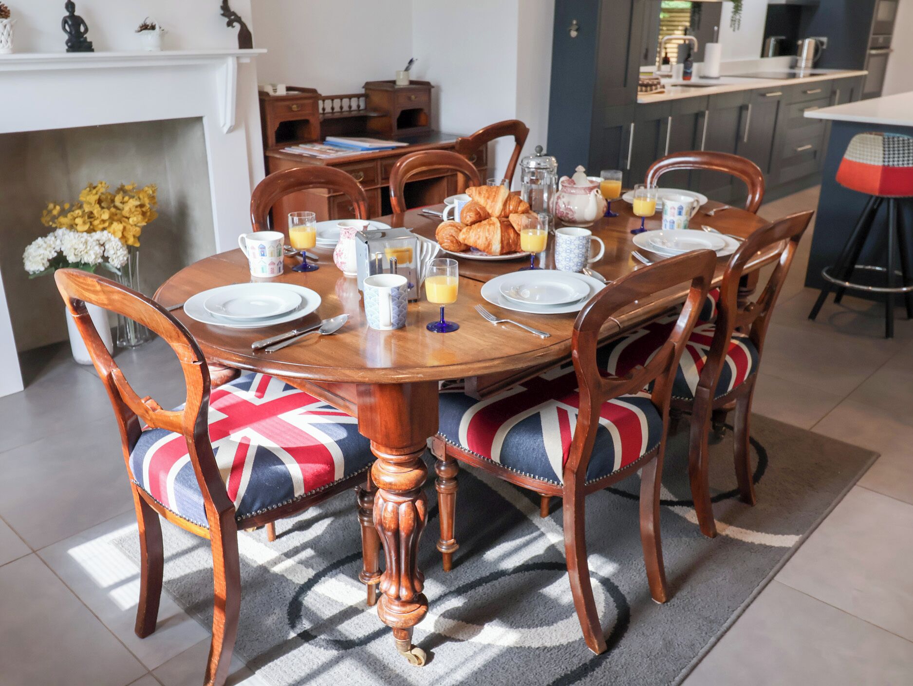Un comedor con una mesa redonda de madera dispuesta para el desayuno, rodeada de sillas tapizadas con cojines con la bandera Union Jack.