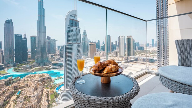 Un balcón con una mesa de mimbre, croissants y vasos de zumo de naranja, con vistas a un paisaje urbano que incluye edificios altos y un puerto deportivo.