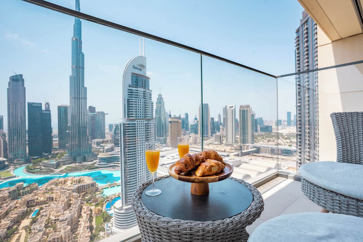 A balcony with a wicker table, croissants, and orange juice glasses, overlooking a cityscape with high-rise buildings and a marina.
