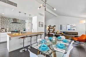 Une cuisine moderne avec un îlot central, un coin repas doté d’une table en verre et un salon avec un canapé et un téléviseur.