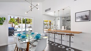 A modern kitchen with a glass dining table, white chairs, and a bar area. - Enchanting Escudero - Luxury PS Ranch Home! (Palm Springs)