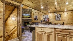 A wooden cabin interior with a stone fireplace, wooden walls, a kitchen with marble countertops, and a dining area.