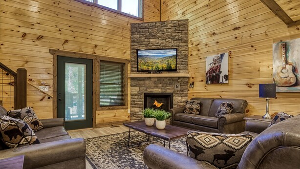 A log cabin interior with a stone fireplace, a television mounted above it, and a cozy seating area with leather sofas and patterned cushions.