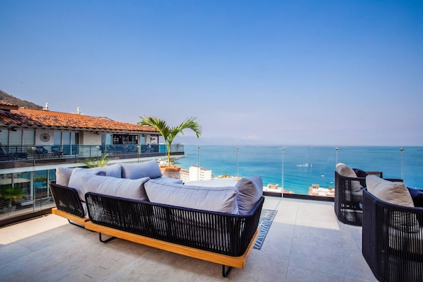 Ein Balkon mit einer Couch und Stühlen mit Blick auf das klare, blaue Meer.