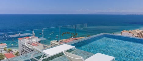 Una piscina infinita en la azotea con agua azul cristalina, con vistas a un puerto deportivo con barcos y una costa lejana.