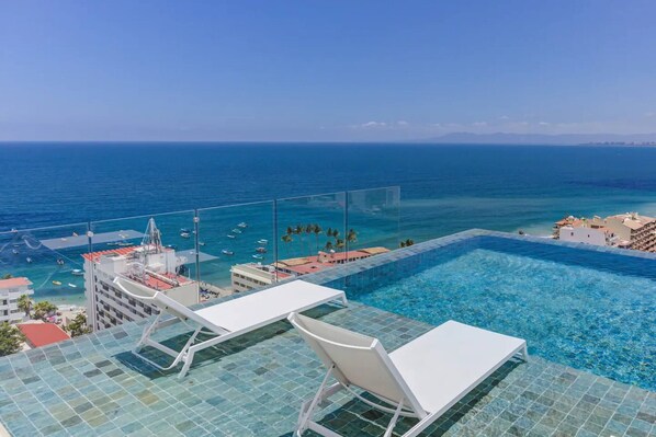 A rooftop infinity pool with clear blue water, overlooking a marina with boats and a distant coastline.
