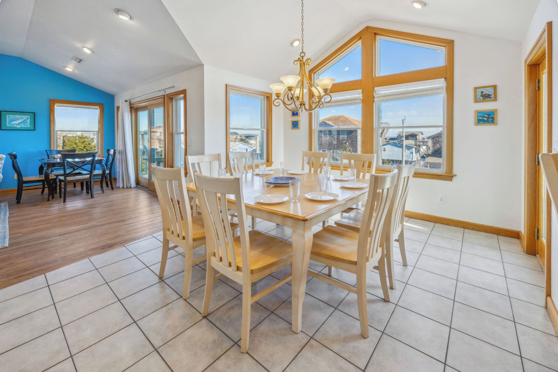 A dining area with a large wooden table and chairs, a chandelier, and large windows offering views of the surrounding area.