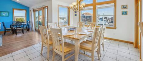A dining area with a large wooden table and chairs, a chandelier, and large windows offering views of the surrounding area.