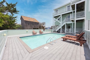 A rooftop pool area with wooden deck, lounge chairs, and a building with a balcony in the background. - Oceanside Dog Friendly w/ Private Pool, Hot Tub, Game Room, Nearby Beach Access (Avon)
