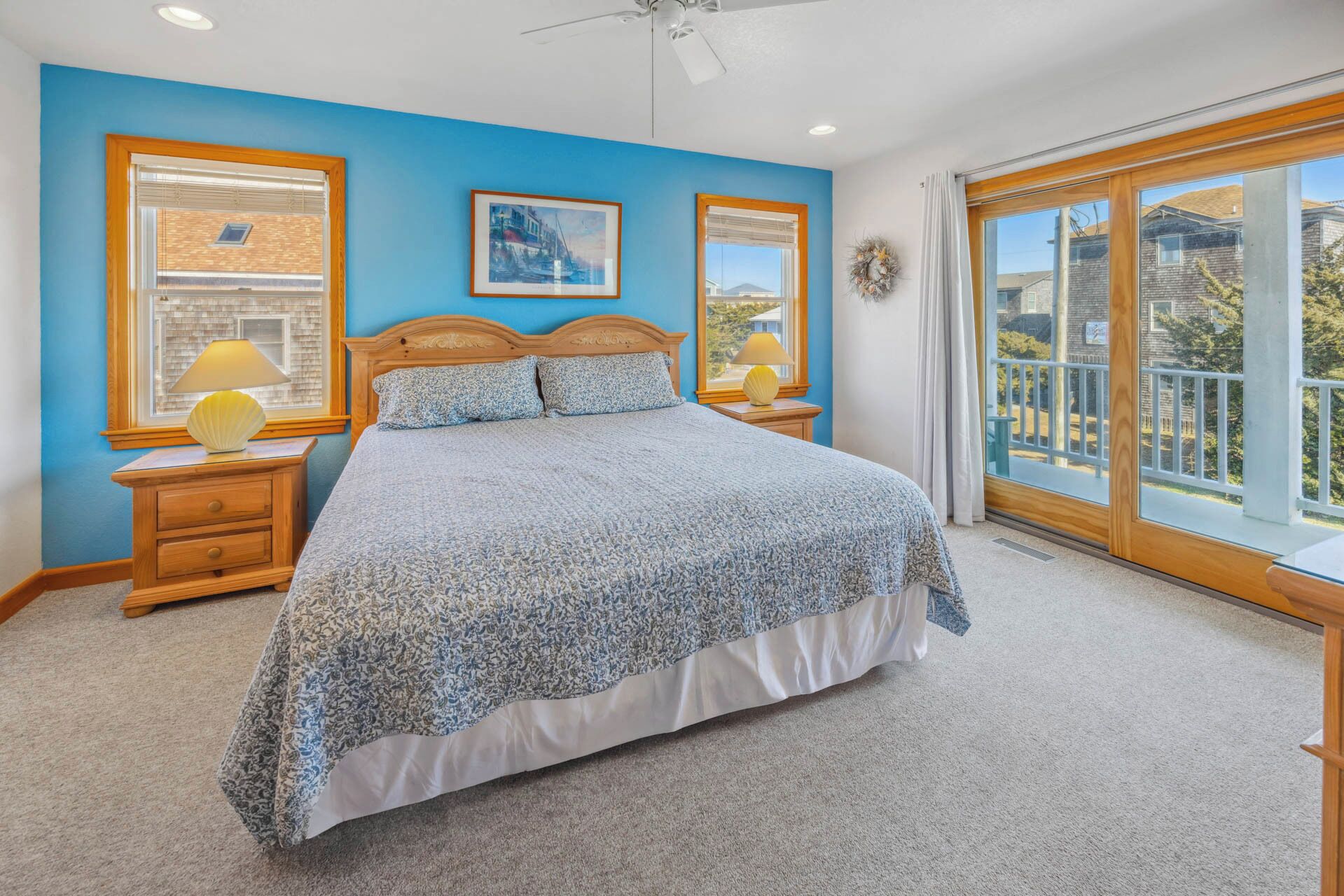 A bedroom with a large bed, two bedside tables with lamps, a ceiling fan, and a view of the outside through a sliding glass door.