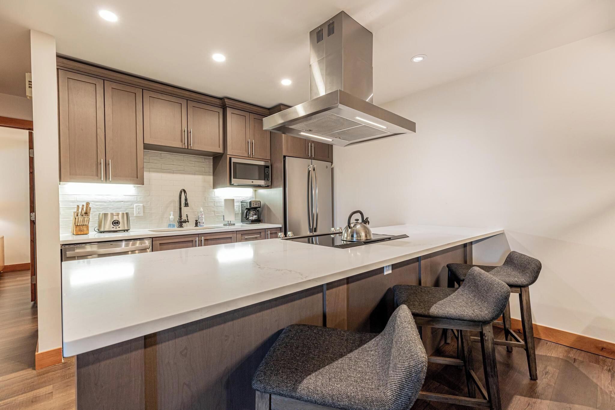 A modern kitchen with wooden cabinets, stainless steel appliances, a large island with bar stools, and recessed ceiling lighting.