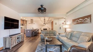 Ein modernes Wohnzimmer mit einem Ecksofa, einem Couchtisch und einem Fernseher auf einer Holzkonsole. Im Hintergrund ist eine Küche mit Essbereich erkennbar.